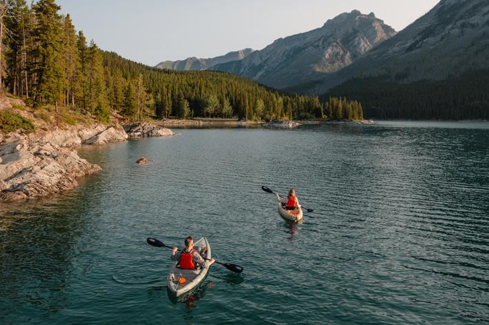Two kayakers paddle on a large lake next to forests and mountains.