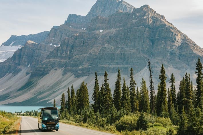 A bus drives next to a forest below a grey mountainside