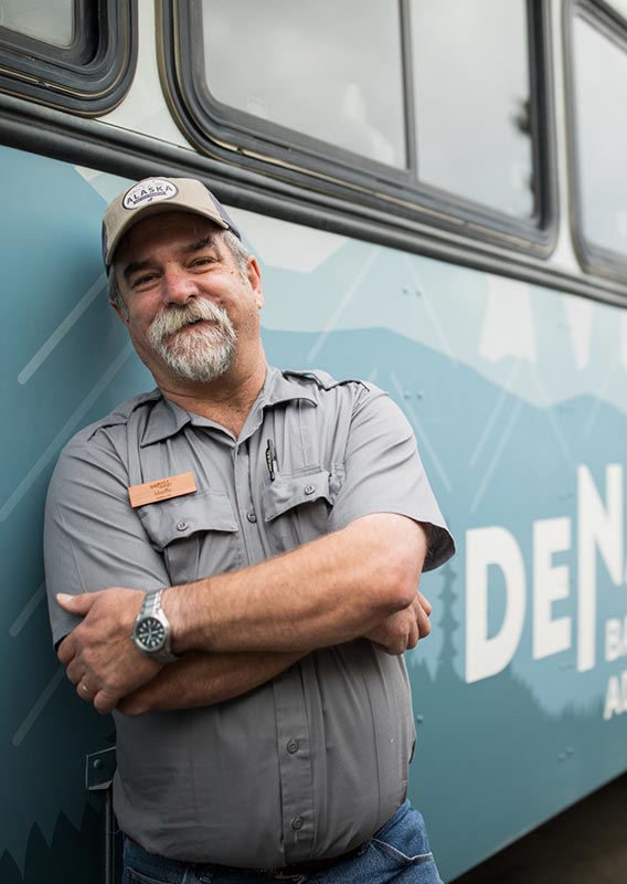 A bus driver leans against a blue Denai Backcountry Adventure bus.