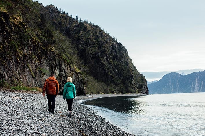 Two people walk along a rocky beach between the ocean and cliffsides.
