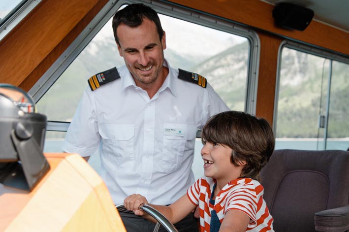 A boat captain stands aside while a child takes the wheel