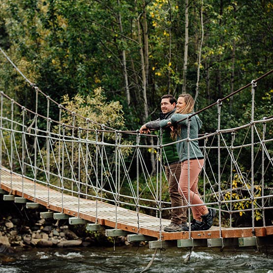 A man and woman stand on a rope bridge suspended over a riverbed.