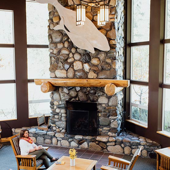A woman sits next to a tall stone fireplace in Windsong Lodge's lobby.