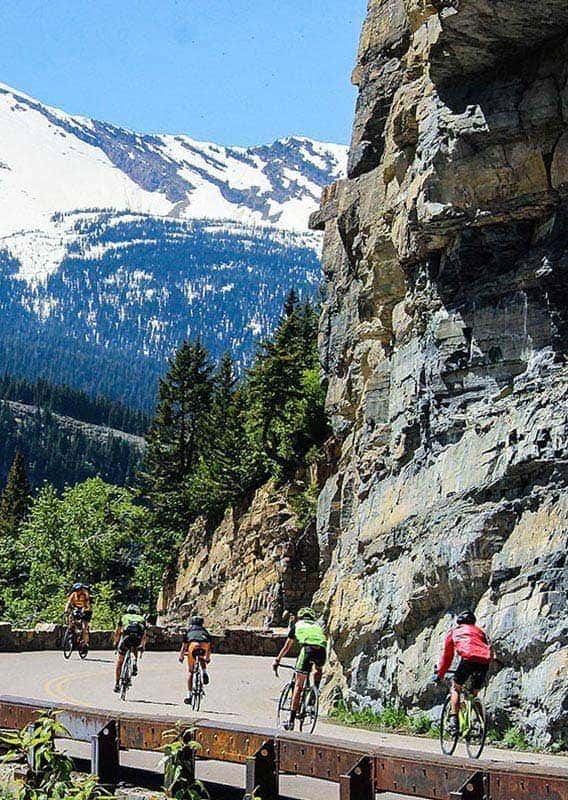 A group of cyclists round a corner alongside rocky cliffs high among mountains.