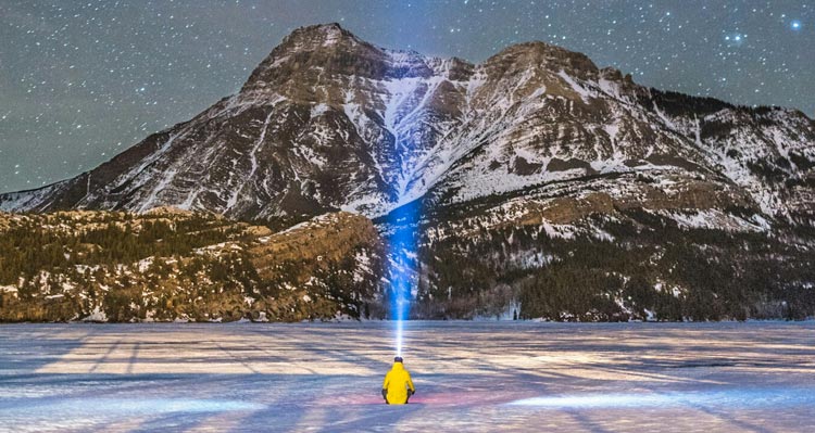 A blue light shines from a man dressed in yellow, sitting on ice below mountains