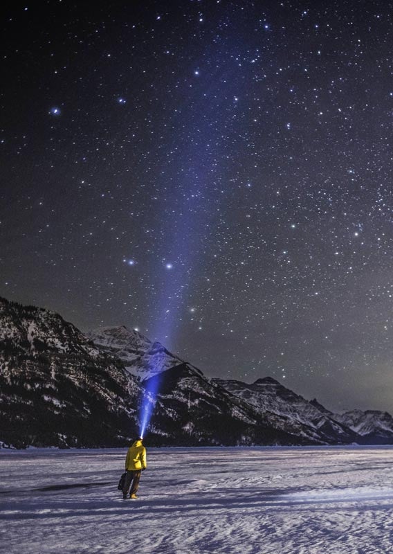 A man in yellow stands on ice, shining his headlamp to the sky.