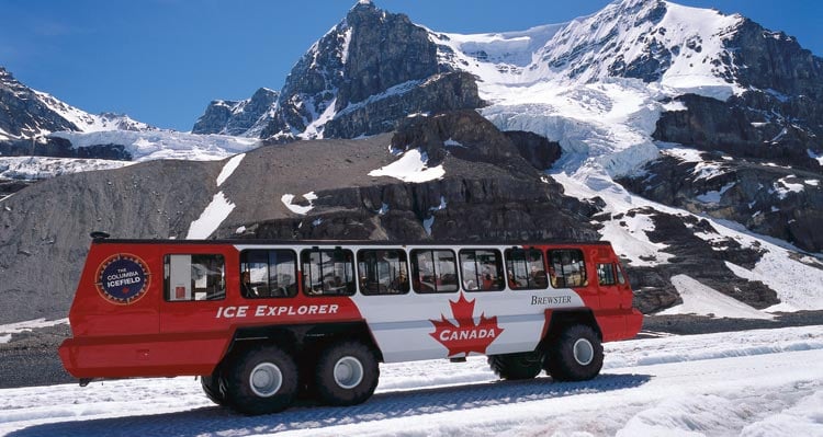 A red and white Ice Explorer bus drives along ice beneath snow-covered mountains.