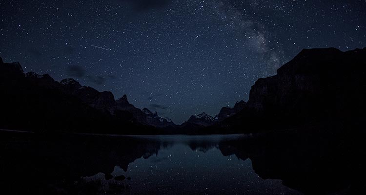 A dark night sky covered in stars above a lake surrounded mountains.
