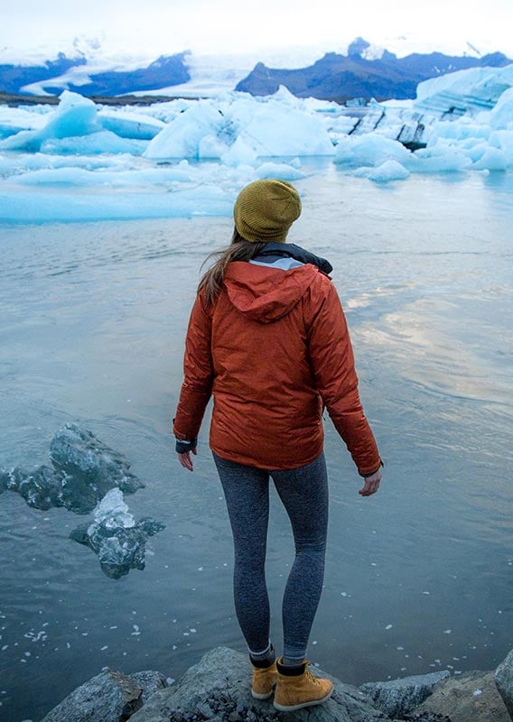 a woman in a red jacket stands at the edge of water with icebergs