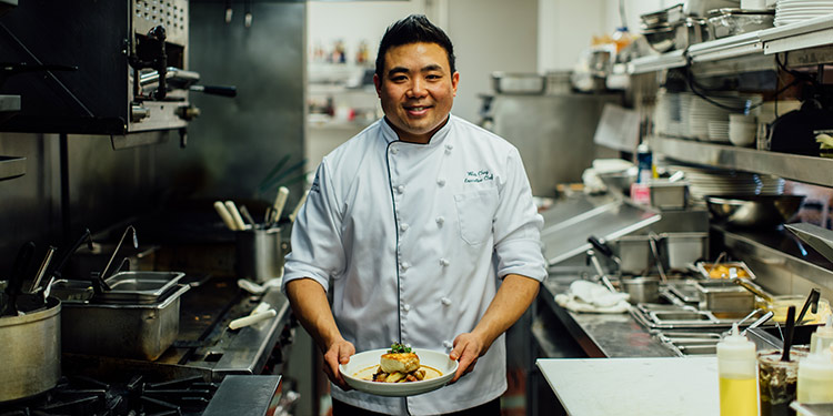 a chef stands in a culinary kitchen holding a plated dish
