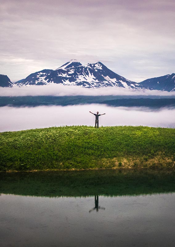A person stands on a green island between some water and fog.