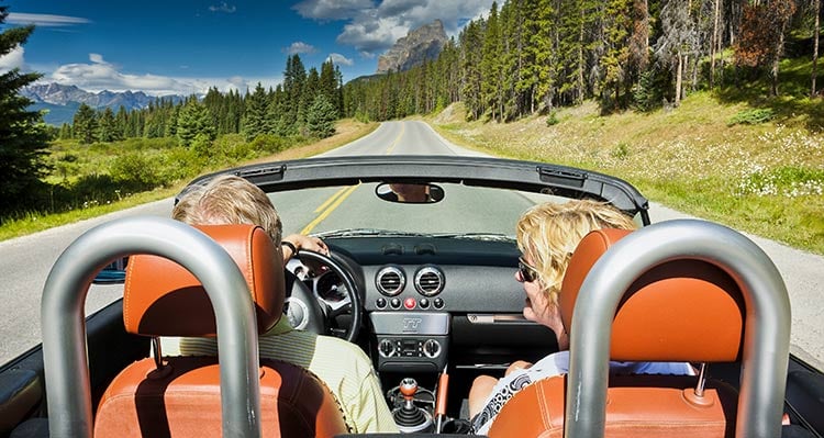 Two people in a convertible driving down an open road toward forests and mountains.