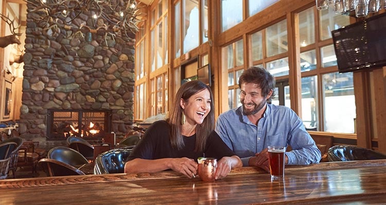 Two people sit at a bar in a dining room with a large stone fireplace.