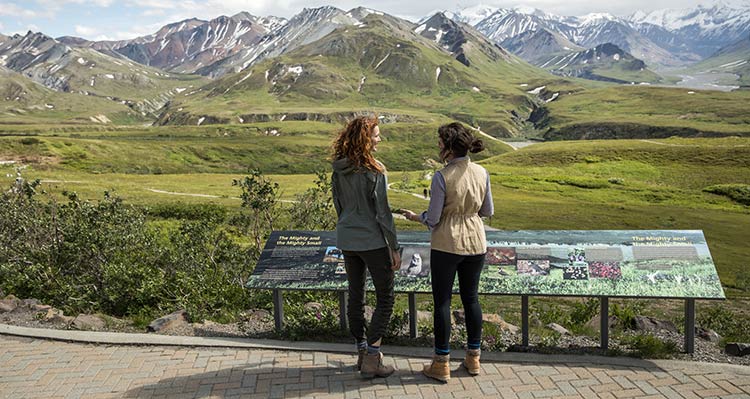 Two people stand at a viewpoint, overlooking tundra and mountains.