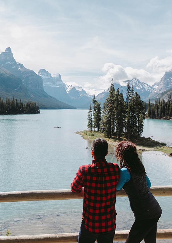 Two people stand overlooking a lake below tall mountains.