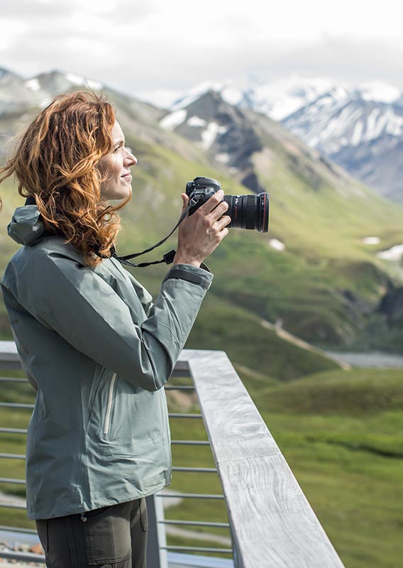 A woman stands at a lookout above tundra, ready to take a photo.