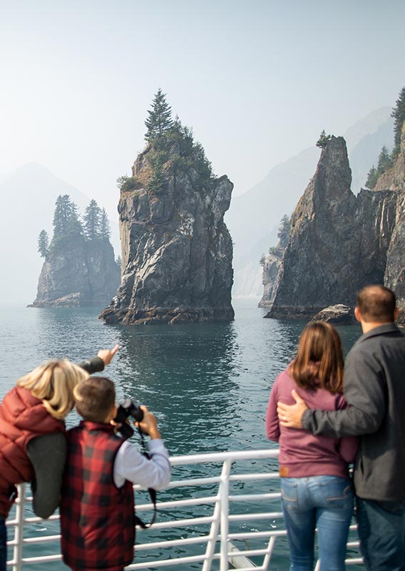 Groups of people stand at the railing of a boat looking towards rock pillars coming from the sea.