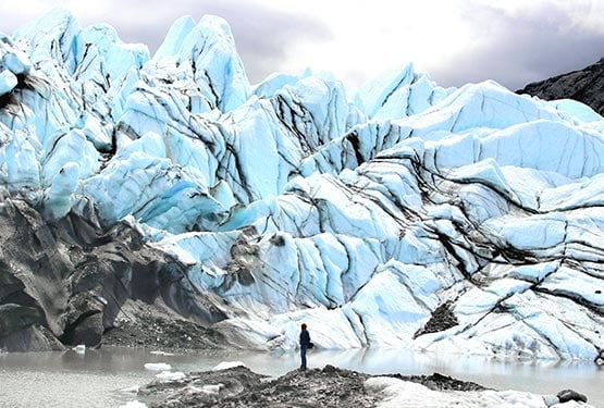 A person stands on a small rocky outcropping at sea, looking at a large glacier.