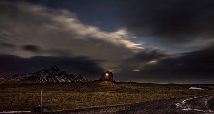 A house in the distance of a dark spooky Icelandic landscape with a single light on.